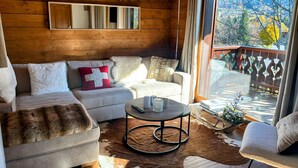 A cozy cabin interior with a wooden ceiling, a sofa with a fur throw, a round coffee table, and a view of a deck with a railing.