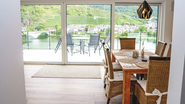 A dining area with a table set for four, a view of a river and bridge, and a large sliding glass door leading to an outdoor seating area.