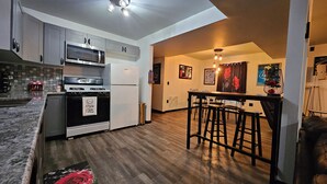 A modern kitchen with a built-in oven, microwave, and refrigerator. There is a wooden dining area with a bar stool and a small table.