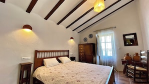 Une chambre à coucher avec un lit en bois, une table de chevet, une commode et un miroir. Il y a deux lampes fixées au mur et une fenêtre avec des rideaux.