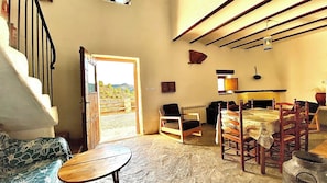 A spacious living room with a large open door leading to an outdoor area, a wooden table, a sofa, and a bed with a wooden headboard. - CASAS RURALES CORTIJO LEONTINO (Taberno)