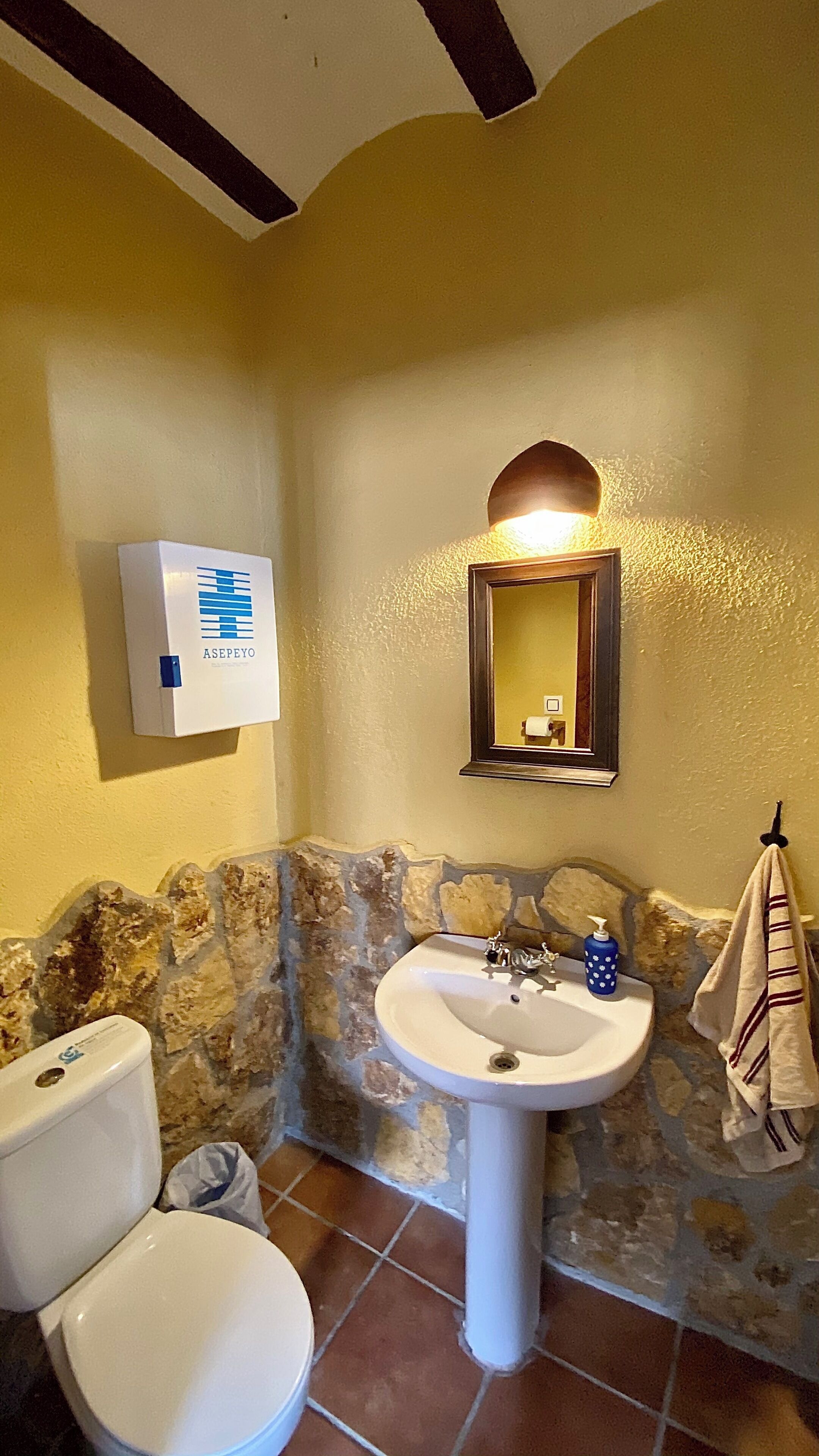A bathroom with a stone wall, a white sink, a toilet, and a mirror with a light above it.