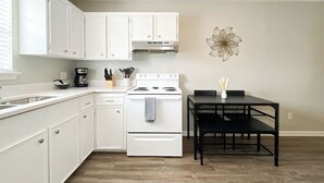 Una cocina con armarios blancos, estufa, cafetera y una mesa de comedor con sillas.