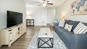 Sala de estar con televisor, mueble de madera para entretenimiento, sofá azul, alfombra estampada y ventilador de techo.