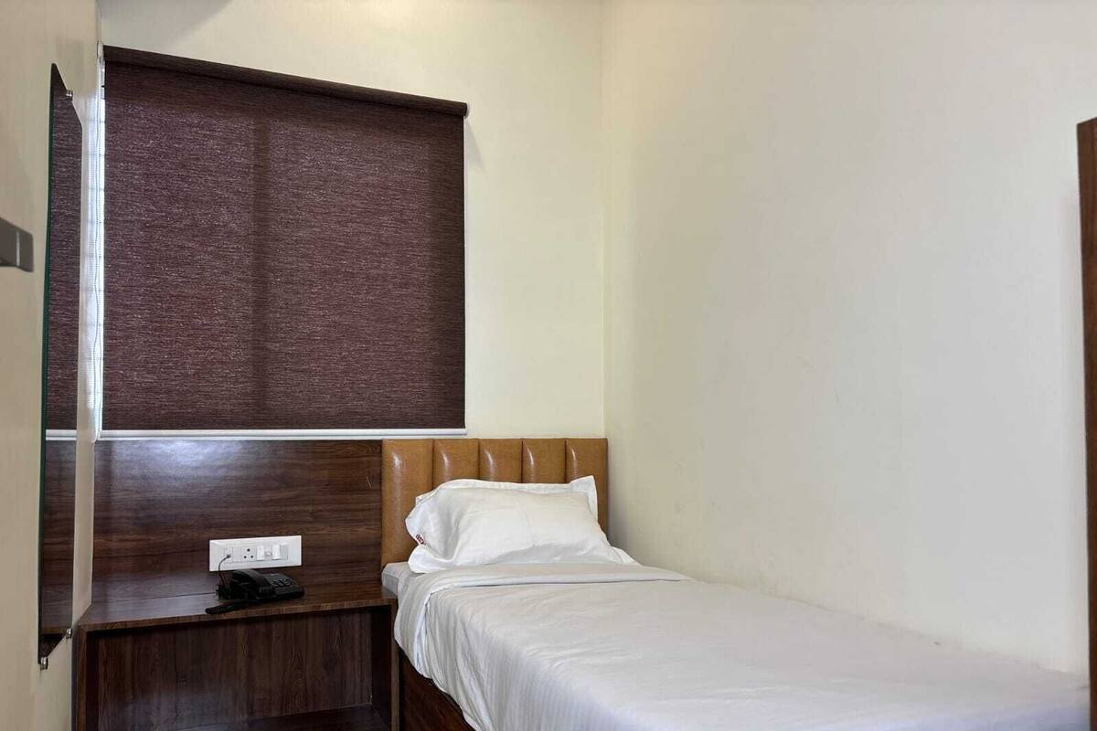 Ein kleines Zimmer mit einem Einzelbett, einem Holz-Kopfteil, einem Nachttisch mit Steckdose und einem braunen Fensterrollo.