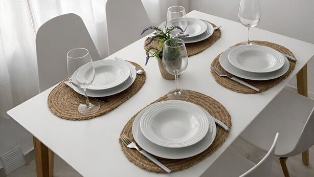 Une table à manger dressée avec de l’assiette blanche, des verres transparents et des sets de table en fibres tressées.