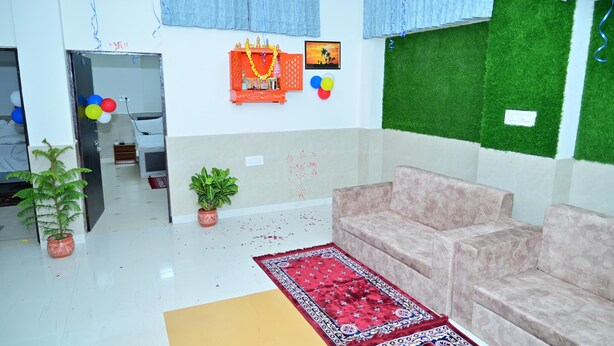 A room with a beige sofa, a red patterned rug, and a green artificial grass wall. There are potted plants and a small altar with decorations. - Hotel Red Inn (Jodhpur)