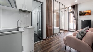 A modern kitchen with a sink and cabinets, a glass door leading to a bathroom, and a living area with a sofa and a painting on the wall. - Lalala Inn Hotel (Ho Chi Minh City)