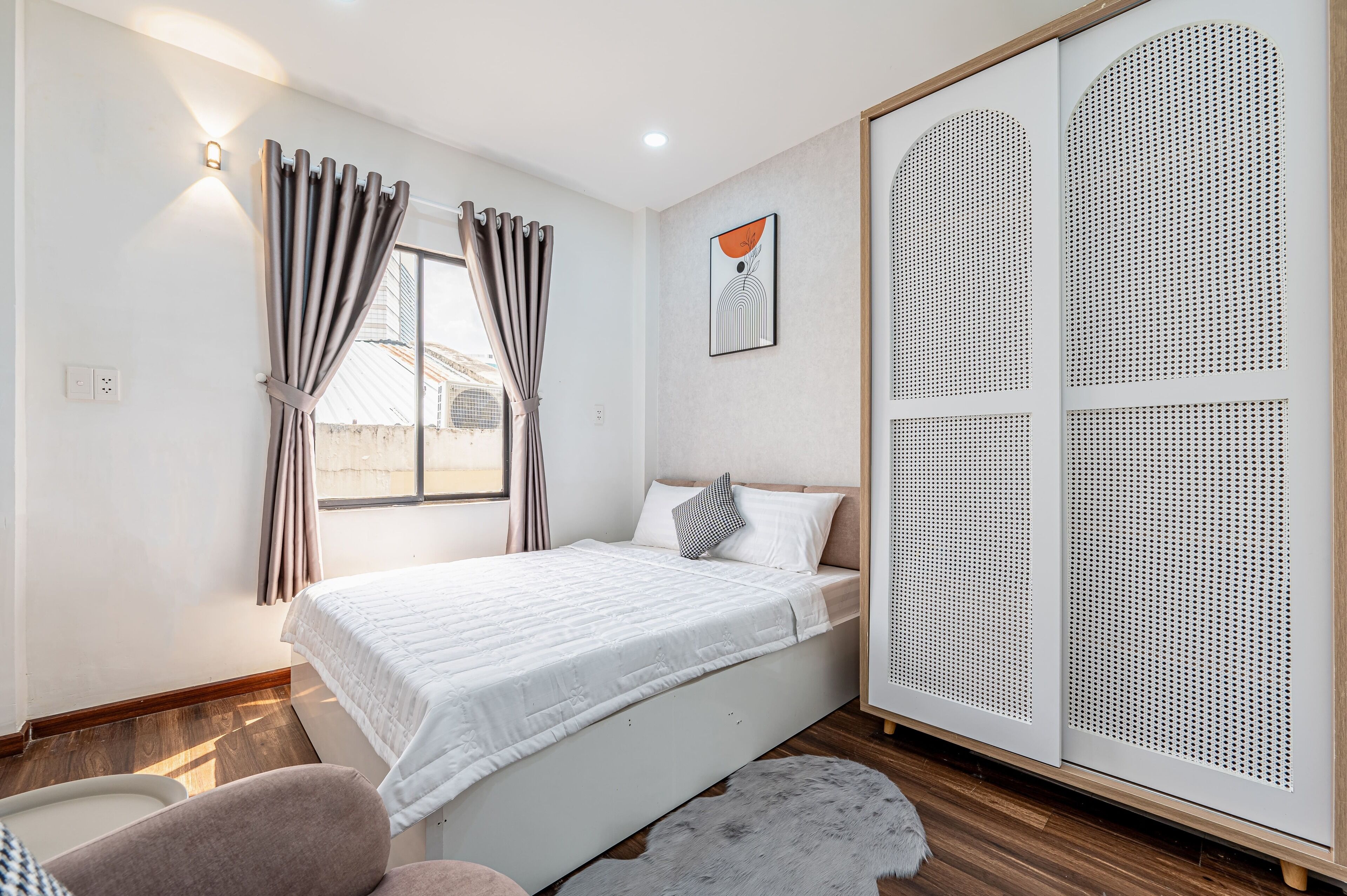 Een slaapkamer met een bed, een kledingkast, een raam met gordijnen, een wandlamp en een vloerkleed.