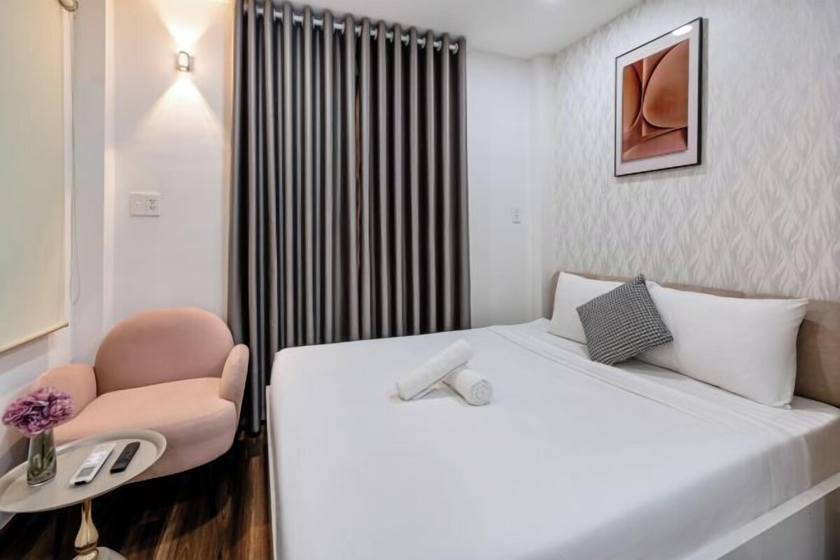 A hotel room with a bed, a chair, a small table with a vase, and a wall-mounted light.