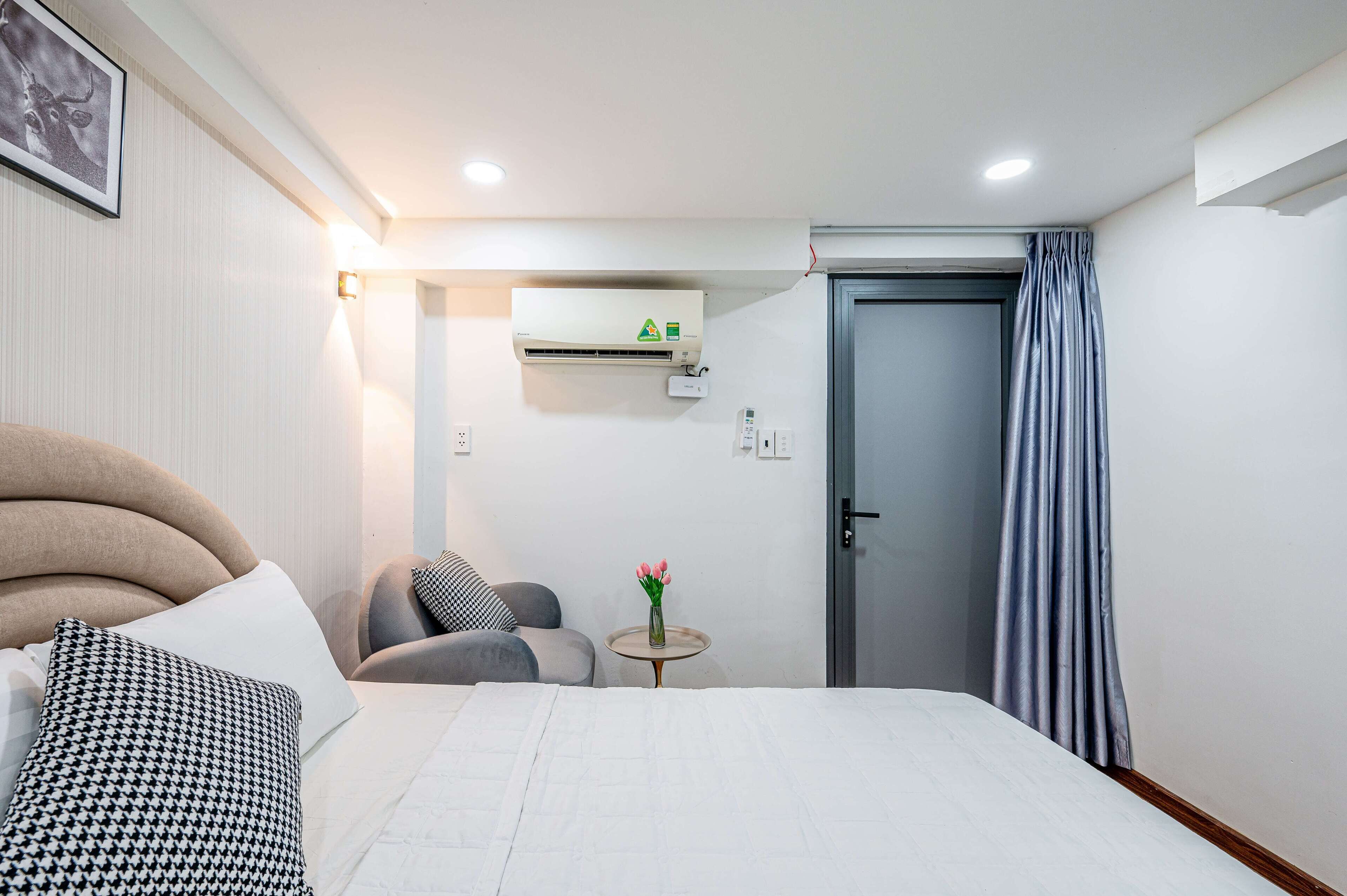 A hotel room with a bed, a chair, a small table with a vase of flowers, an air conditioning unit, and a door with curtains.