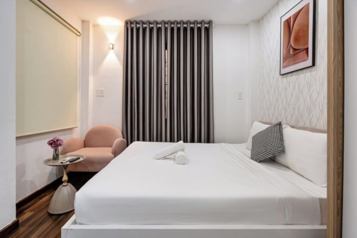 Quarto de hotel com uma cama grande, uma mesa de cabeceira com abajur, uma cadeira, uma janela com persianas e uma obra de arte na parede.