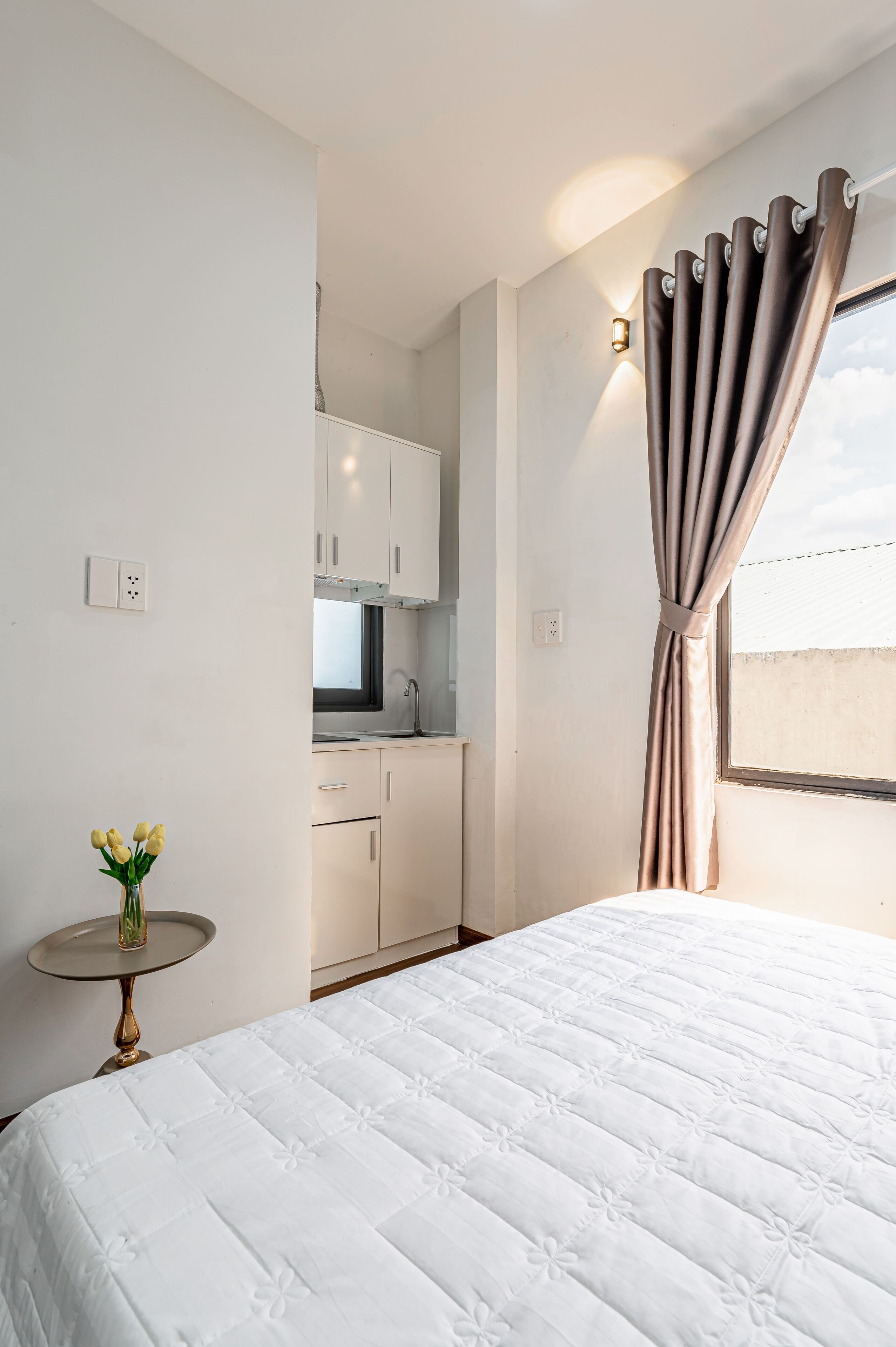 A hotel room with a bed, a small table with a vase, a kitchenette, and a window with curtains.