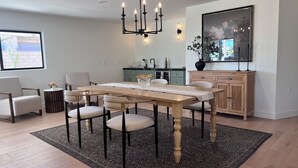 A dining area with a wooden table, chairs, a chandelier, a kitchen in the background, and a large window with a view.