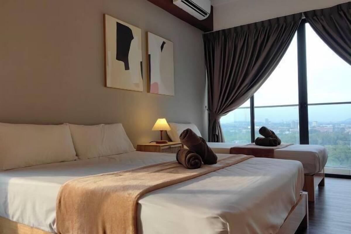 A hotel room with two beds, a large window with curtains, a lamp on a bedside table, and artwork on the wall.