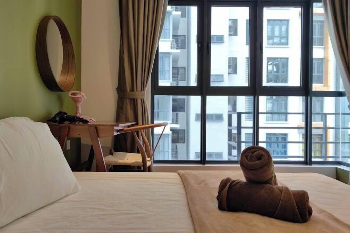 A hotel room with a bed, a desk, a chair, a large window, and a view of buildings outside.