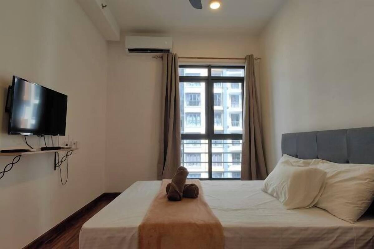 A hotel room with a bed, a flat-screen TV mounted on the wall, a window with curtains, and an air conditioning unit.