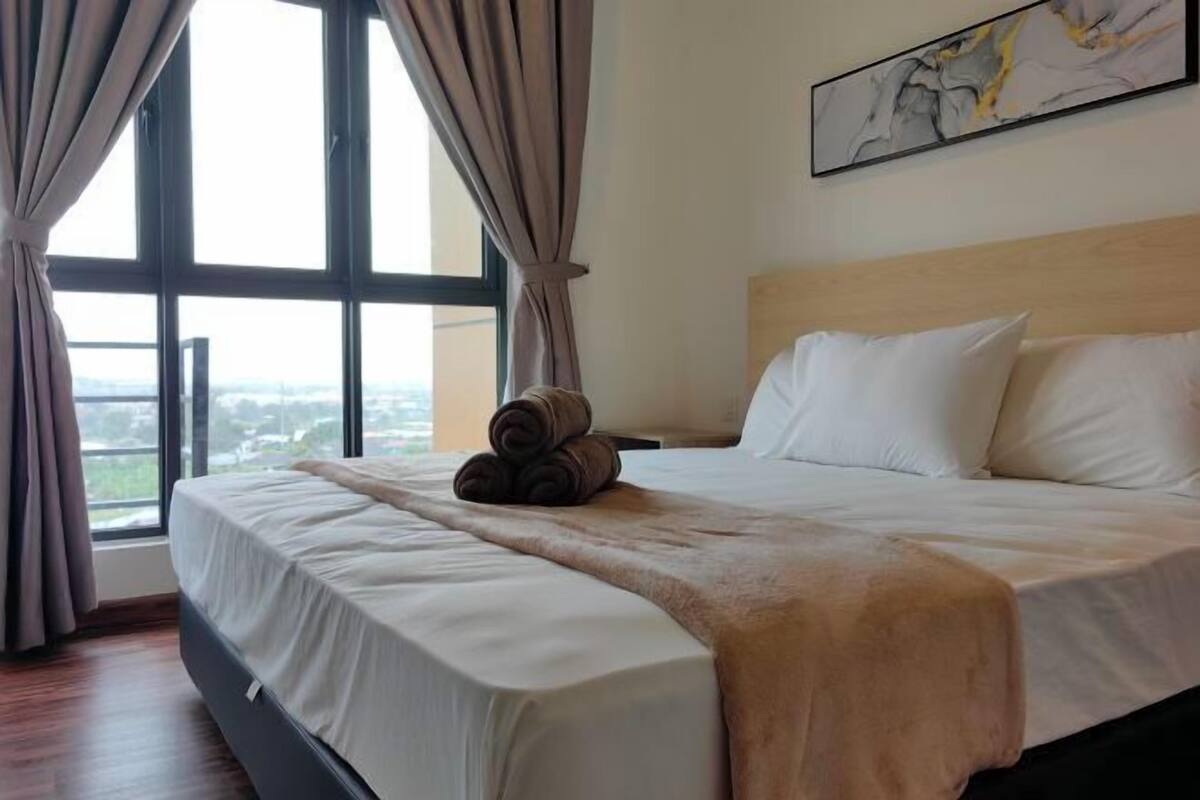 A hotel room with a large window, a bed with white linens, a headboard, and a painting on the wall.