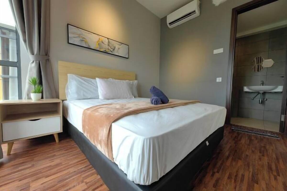 A modern hotel room with a large bed, a bedside table, a nightstand, a window with curtains, an air conditioning unit, and a bathroom with a sink and shower.