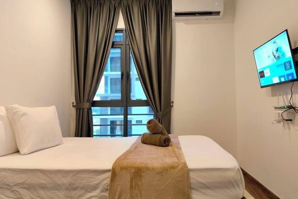 A hotel room with a bed, a TV on the wall, a window with curtains, and a view of buildings outside.