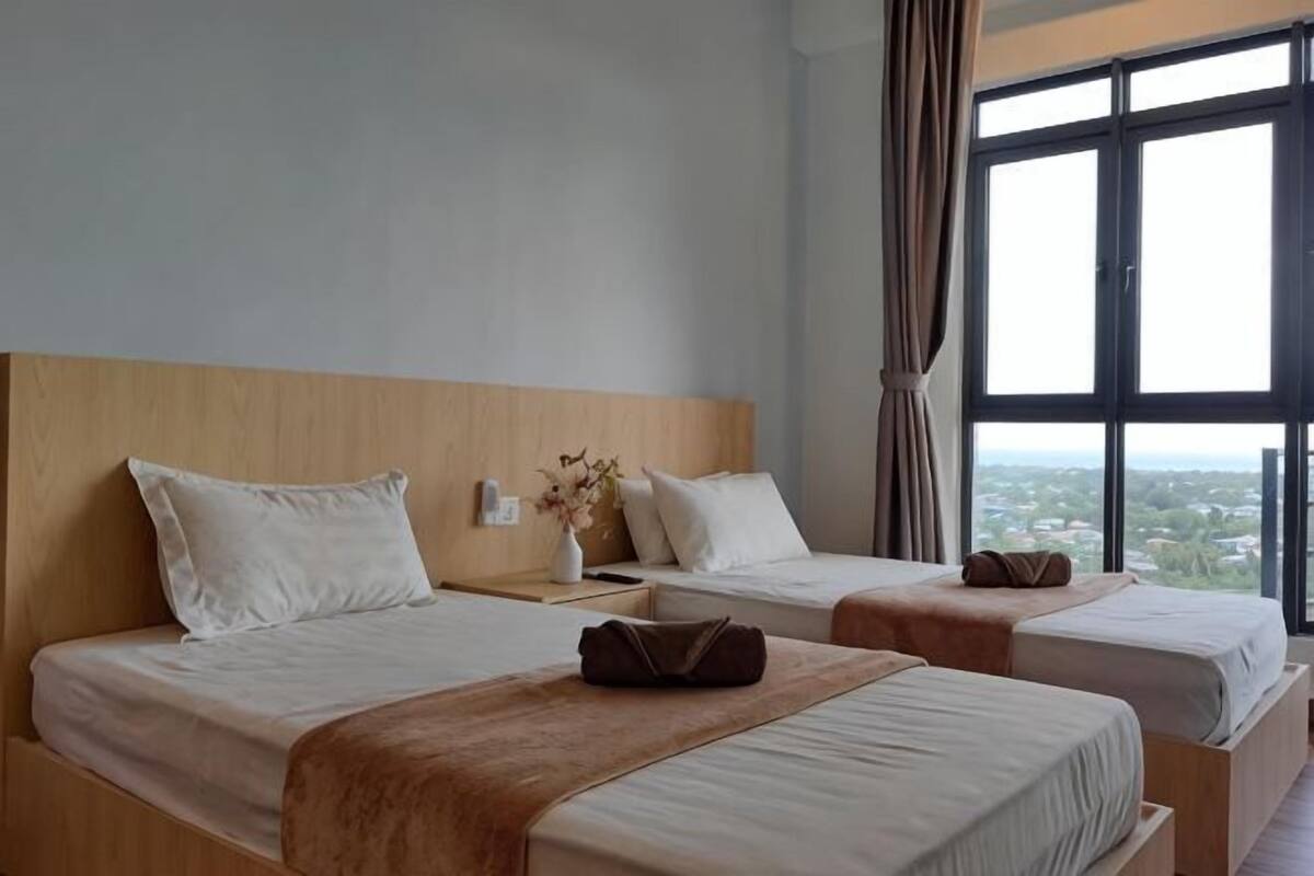 Two single beds with wooden headboards, a nightstand with a vase, and a large window with curtains.