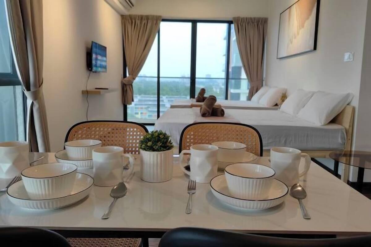 A hotel room with a dining table set with white bowls, plates, and cutlery, a potted plant, and a bed with pillows.