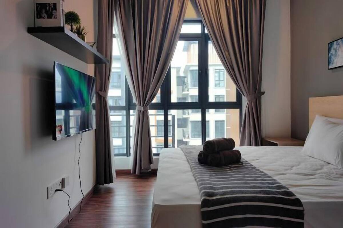 A hotel room with a bed, a TV mounted on the wall, a window with curtains, and a view of buildings outside.