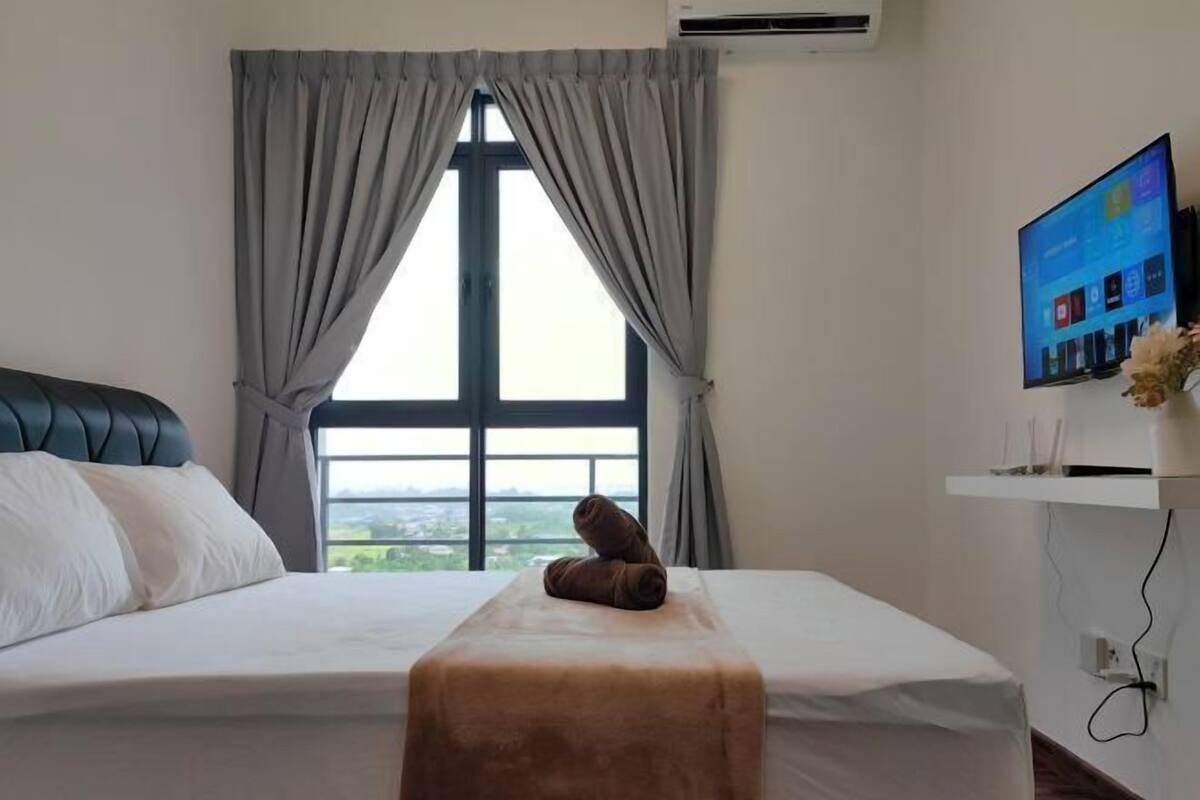 A hotel room with a bed, a window with curtains, a flat-screen TV on a stand, and a view of the outdoors.