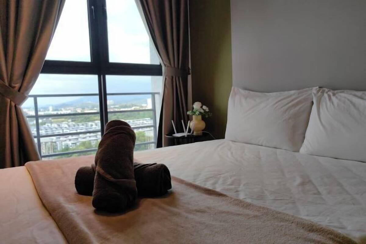 A hotel room with a bed, a window with curtains, a small table with a vase, and a view of the cityscape.