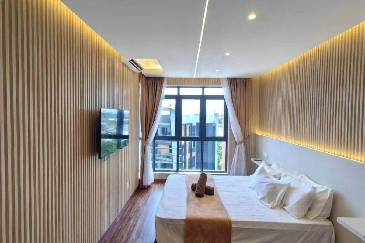 A modern hotel room with a large bed, a flat-screen TV mounted on the wall, and a window providing a view of the cityscape.
