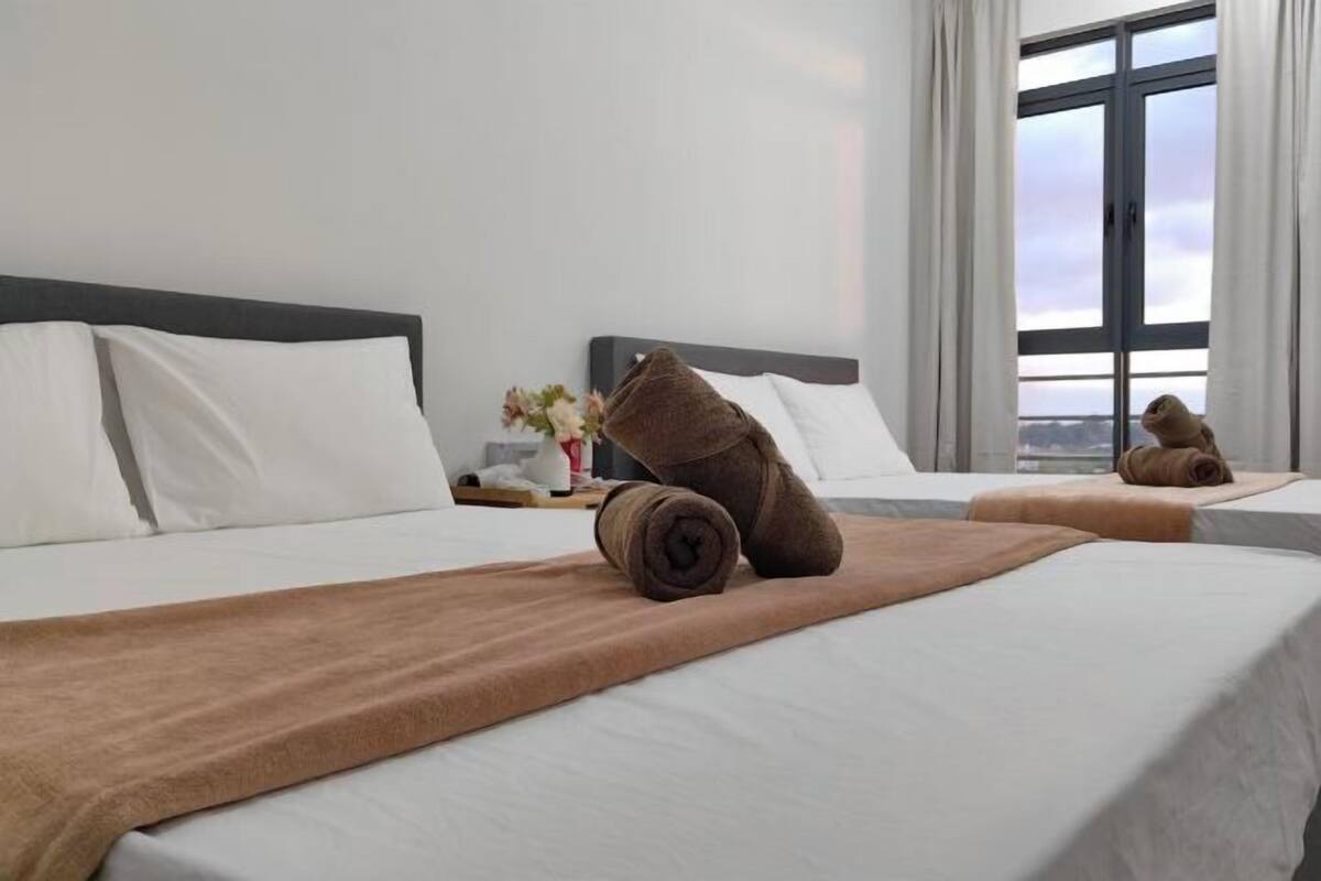 A hotel room with two beds, white linens, brown towels, a nightstand with a vase of flowers, and a large window with curtains.