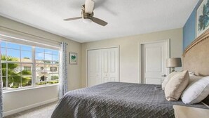 Un dormitorio con cama, ventilador de techo, ventana con cortinas y vista a un edificio.