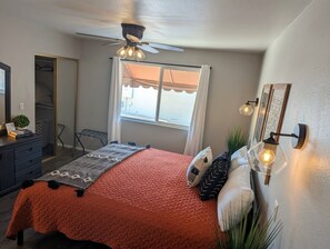 Un dormitorio con cama, ventilador de techo, ventana con cortinas, mesita de noche y lámpara de pared.