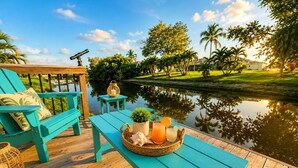Een houten terras met een turquoise tafel en stoelen, een klein tafeltje met een mand drankjes, en uitzicht op een rustig wateroppervlak en groen.