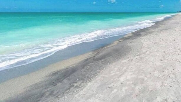 Een strand met helder turquoise water en een zandstrand, onder een blauwe lucht met losse wolken.