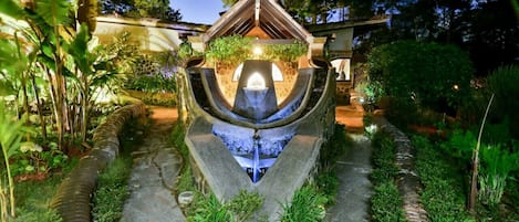 A stone water feature with a central fountain, surrounded by greenery and a pathway.