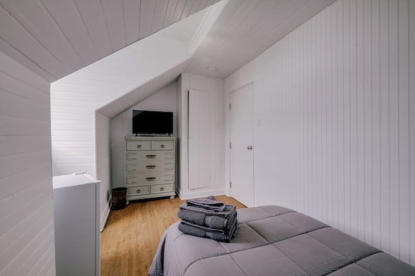 Een slaapkamer met een schuine plafond, een bed met grijze beddengoed, een dressoir met lades en een televisie aan de muur.