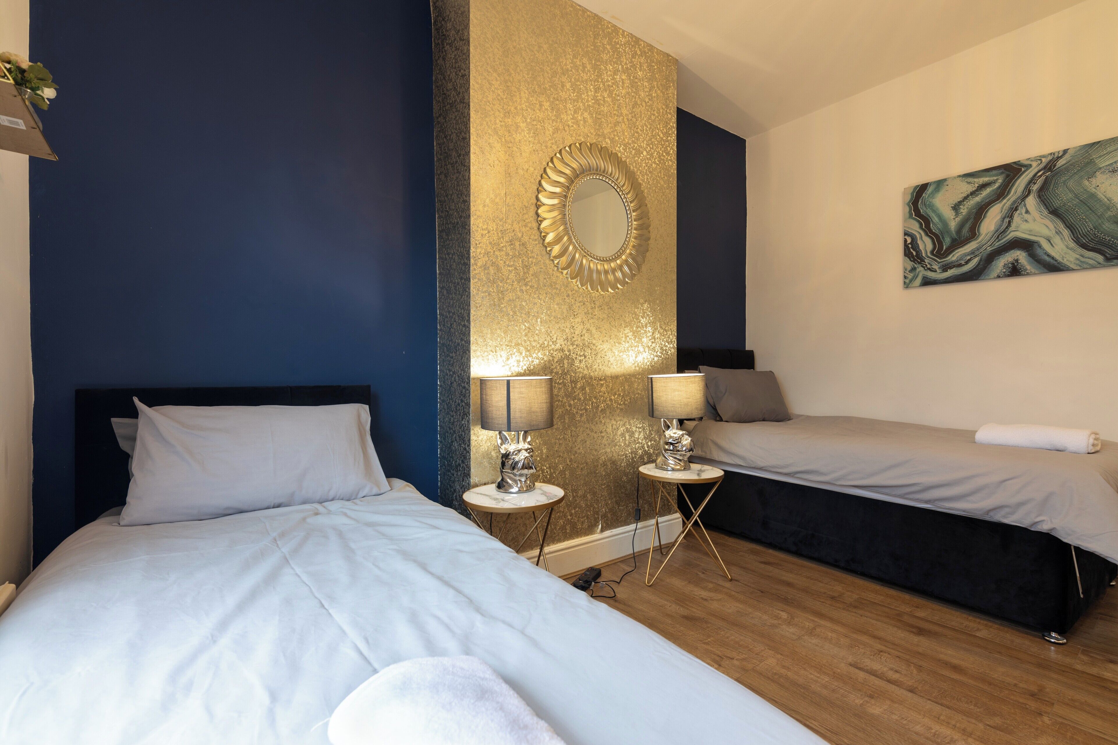 A hotel room with two beds, a gold accent wall, a marble-like artwork, and a small table with lamps.