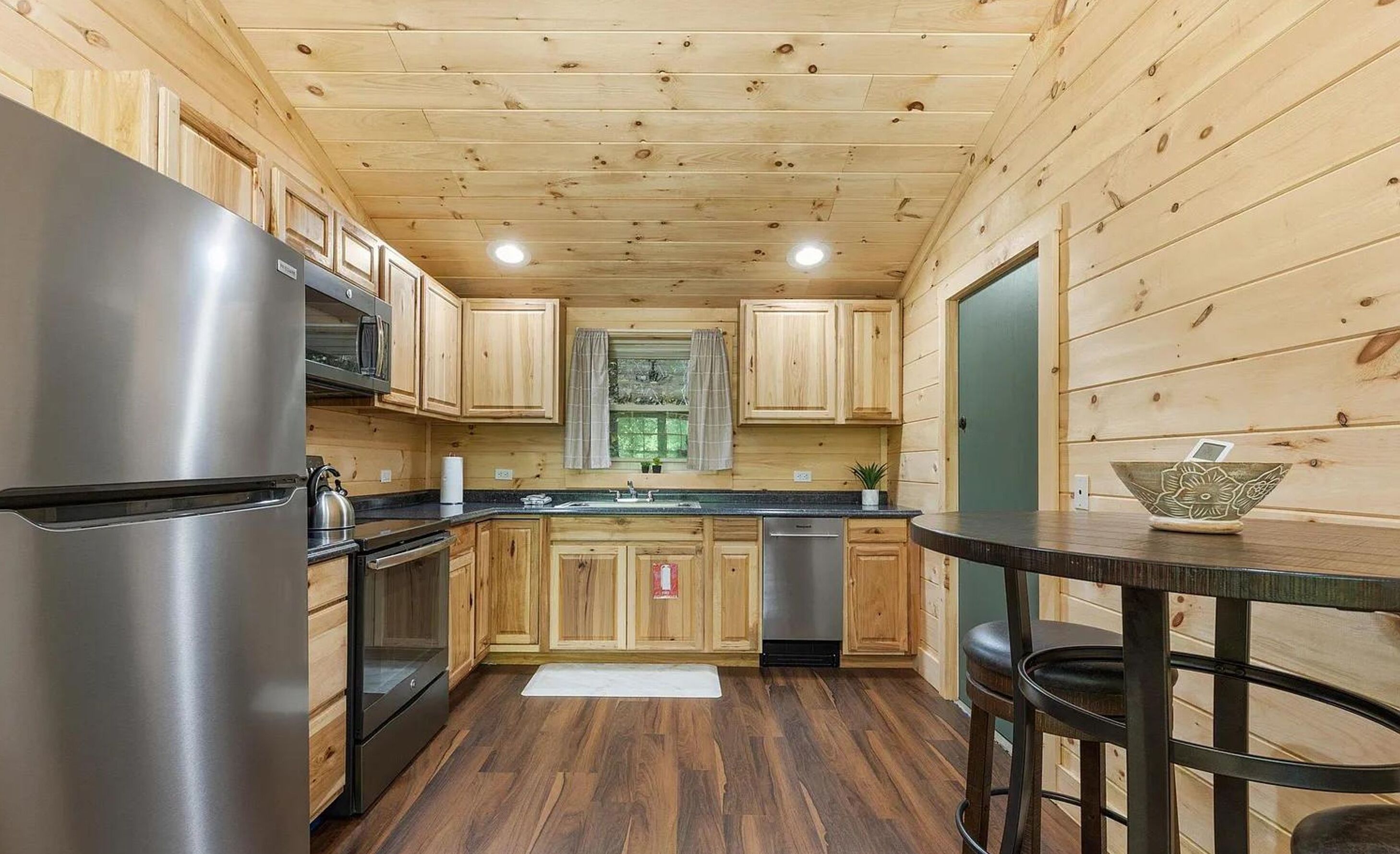 A compact kitchen with wooden walls and a central island, equipped with stainless steel appliances and a small window.