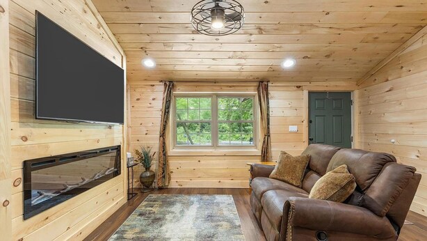 Ein gemütliches Blockhauss-Interieur mit Holzvertäfelung, einem Ledersofa, einem Flachbildfernseher und einem Fenster mit Vorhängen.