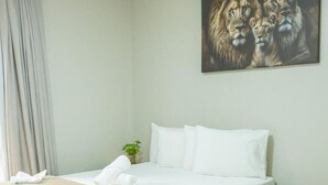 Una habitación de hotel con una cama grande, una cama más pequeña, un cuadro de leones en la pared, una mesita de noche y una ventana con cortinas.