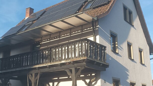 Een huis met twee verdiepingen, een balkon, zonnepanelen op het dak en een stenen muur ervoor.