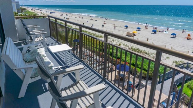 Een balkon met witte stoelen en uitzicht op een strand met parasols en mensen.