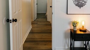 A hallway with wooden flooring, white walls, and a door leading to another room. There is a framed pinecone artwork on the wall and a small table with a lamp and flowers. - "Morning View 2" Cozy Modern Stay - College/Hiking (Berea)