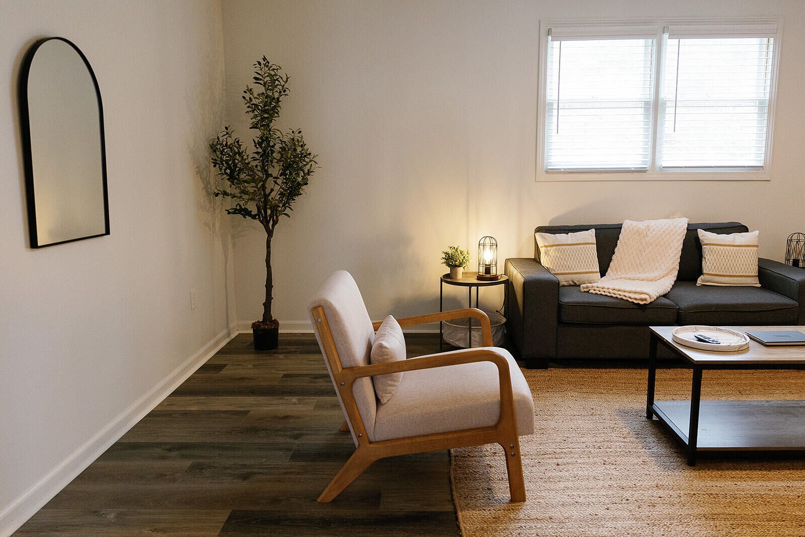 Uma sala de estar com piso de madeira, um sofá, uma poltrona, uma mesa de centro, um abajur e um vaso com planta.