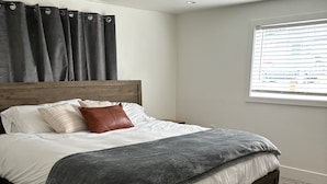 A bedroom with a wooden bed, grey bedding, a brown pillow, and a window with blinds. - Charming 3-bedroom house with WiFi, AC , TESLA Charger in tranquil Lincoln City (Lincoln City)
