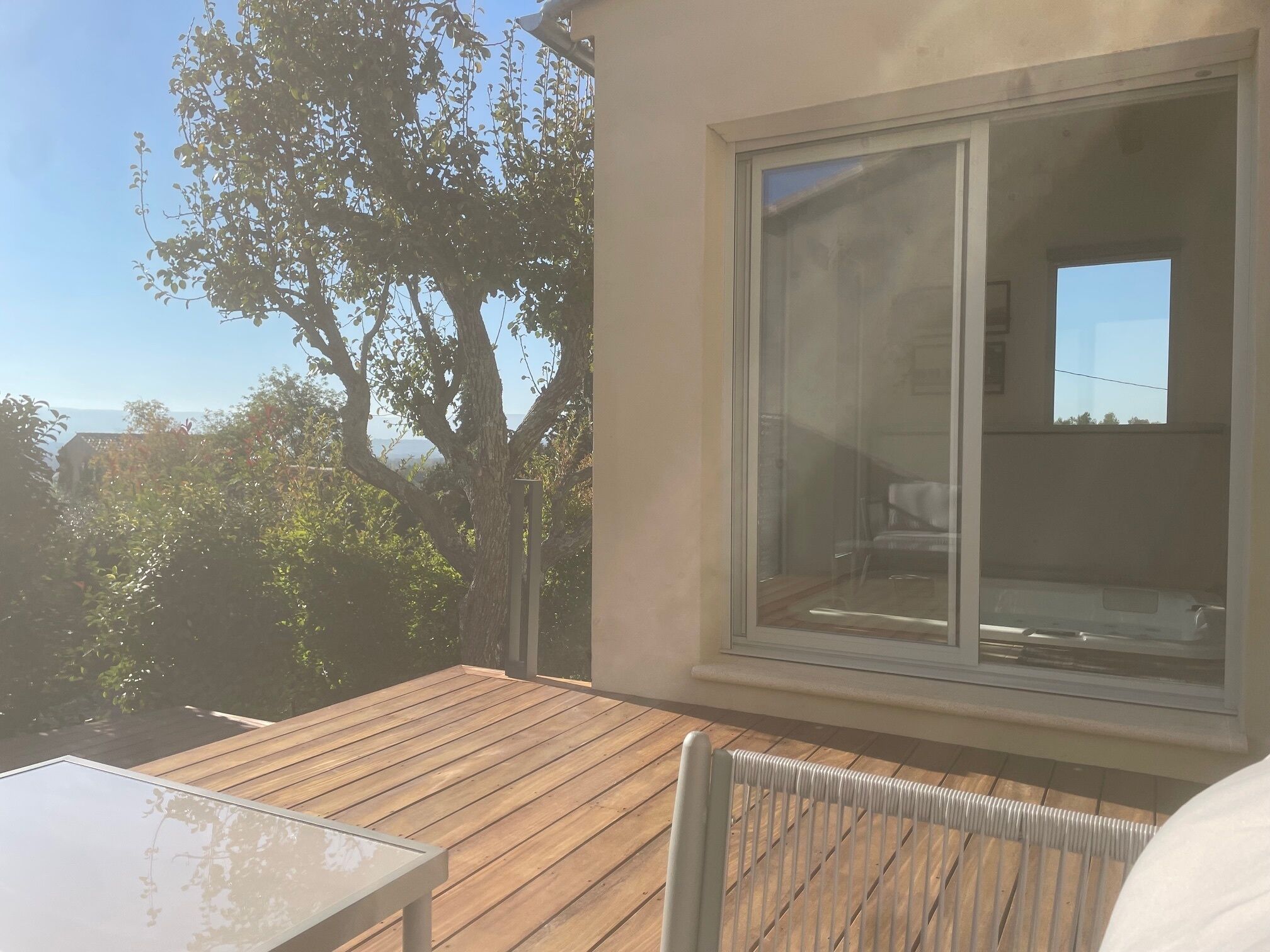Eine Holzterrasse mit einem Glastisch und einem Stuhl, ein großes Fenster und ein Baum im Hintergrund.