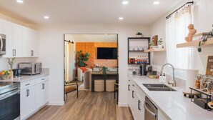 Een moderne keuken met witte kasten, een magnetron en een spoelbak. Er is een eethoek met een tafel en stoelen, en een tv aan de muur bevestigd.