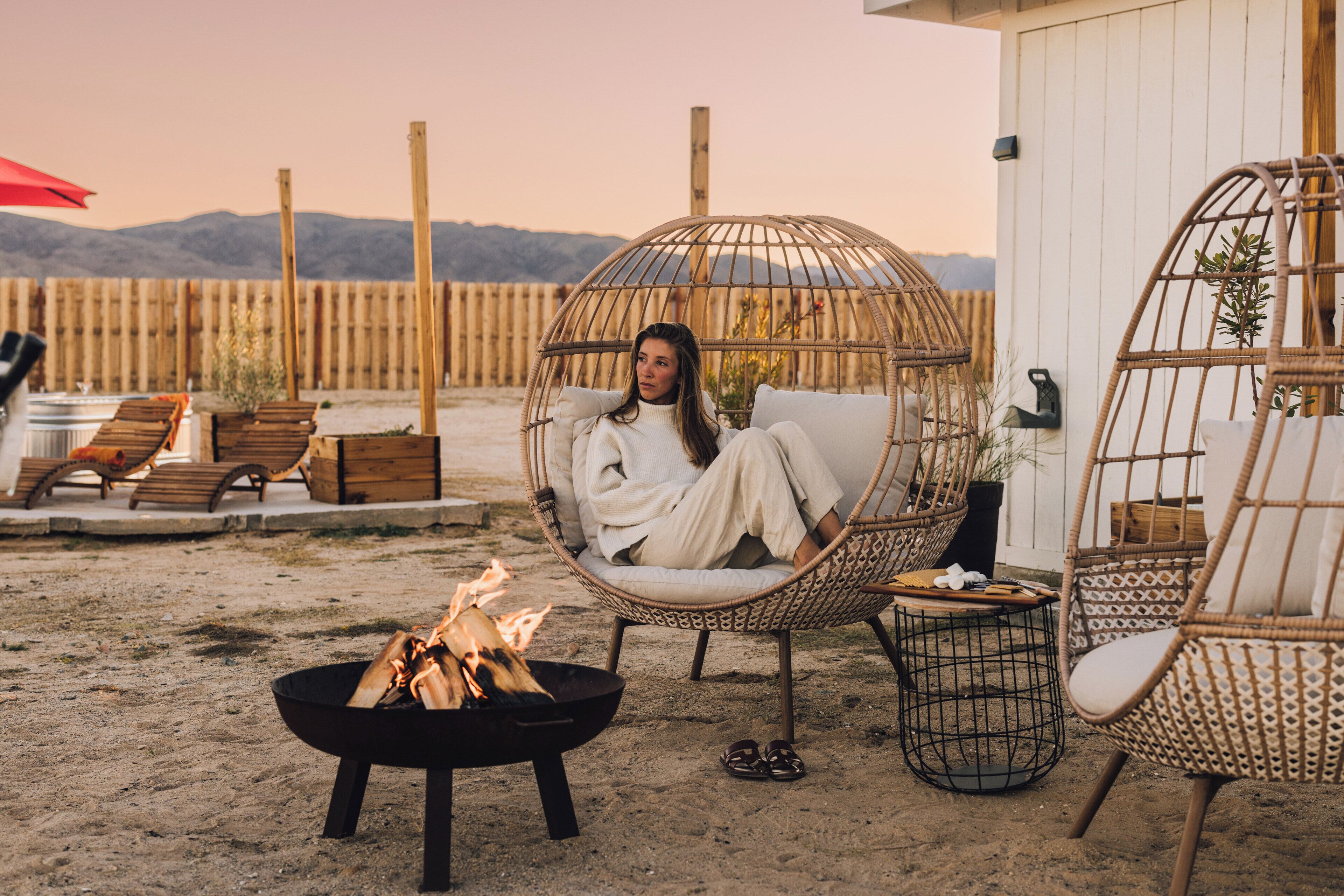 Une personne se détend dans un fauteuil en osier près d’un foyer sur une plage de sable, avec des chaises longues en bois et un bâtiment blanc en arrière-plan.