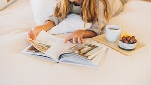 Iemand leest een boek in bed, met een kop koffie en een schaal fruit.